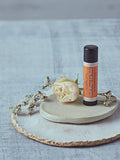 Sweet Orange Lip Balm tube standing on a ceramic dish with a white rose