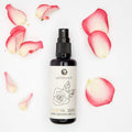 Flat lay of Damask Rosewater spray bottle surrounded by pink rose petals on a white background